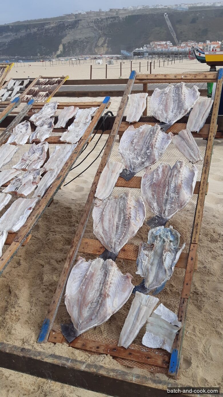 Poisson en train de sécher sur les paneiros de la plage de Nazaré, la falaise du Sítio et son funiculaire au fond, une barque traditionnelle marquée NAZARÉ